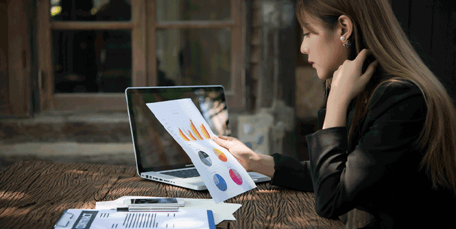 Mulher sentada em frente a um notebook e analisando um papel com dados